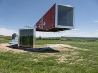 a shipping container is suspended from the side with its doors open on top of it, in front of a grassy field with a large blue sky background