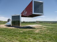 a shipping container is suspended from the side with its doors open on top of it, in front of a grassy field with a large blue sky background