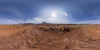 an open desert with red dirt and dirt fields and clouds overhead with a mountain and blue sky