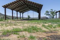 an open covered shelter next to a dirt field with green grass and trees in the background