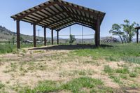 an open covered shelter next to a dirt field with green grass and trees in the background