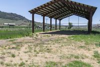 an open covered shelter next to a dirt field with green grass and trees in the background