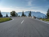 an empty open road with trees, grass and mountains in the distance on a sunny day