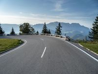 an empty open road with trees, grass and mountains in the distance on a sunny day