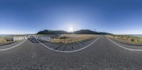 a panorama lens view of an open road with the sun in the distance and mountains in the background