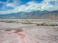 a sandy area with a few pink rocks in it and mountains in the background and a blue sky and some white clouds in the distance