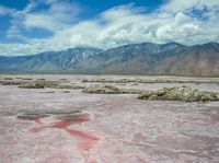 a sandy area with a few pink rocks in it and mountains in the background and a blue sky and some white clouds in the distance