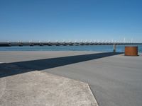 a concrete bench on the sidewalk near water with sailboats in distance behind it,