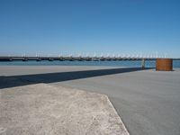 a concrete bench on the sidewalk near water with sailboats in distance behind it,