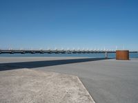 a concrete bench on the sidewalk near water with sailboats in distance behind it,