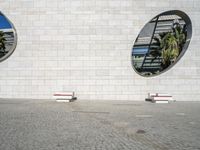 two round mirrors and two benches sit on a block surface near buildings, trees, and a building with windows