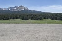 a dirt field and road near pine trees and mountain tops against blue sky with wispy clouds