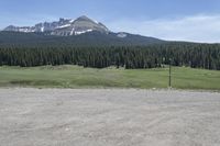 a dirt field and road near pine trees and mountain tops against blue sky with wispy clouds