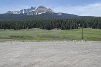 a dirt field and road near pine trees and mountain tops against blue sky with wispy clouds