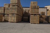 large group of wooden pallets stacked up outside of a warehouse building for sale, with some boxes and a man with a fork in handrail in the background