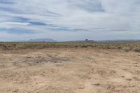 a dirt field on an empty plain with mountains in the distance under clouds and blue sky