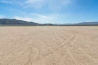 a sandy area with two mountains and sparse grass on the horizon in the distance is dirt that runs through the foreground