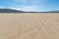 a sandy area with two mountains and sparse grass on the horizon in the distance is dirt that runs through the foreground