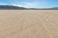 a sandy area with two mountains and sparse grass on the horizon in the distance is dirt that runs through the foreground