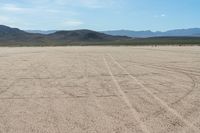 a sandy area with two mountains and sparse grass on the horizon in the distance is dirt that runs through the foreground