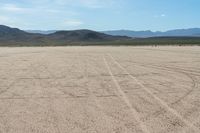 a sandy area with two mountains and sparse grass on the horizon in the distance is dirt that runs through the foreground