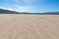 a sandy area with two mountains and sparse grass on the horizon in the distance is dirt that runs through the foreground