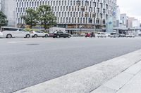 a car driving down a city street past a tall building, with many cars parked along it