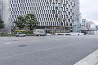 a car driving down a city street past a tall building, with many cars parked along it