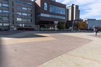 two buildings with a person skateboarding across the sidewalk from one another while another sits on a bicycle in the background