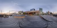 an oval shaped wooden structure at the waterfront of a city with some buildings in the background