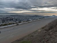 the sky is overcast with a big city in the distance as people walk on the side of a long hill