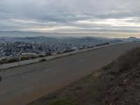 the sky is overcast with a big city in the distance as people walk on the side of a long hill