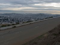 the sky is overcast with a big city in the distance as people walk on the side of a long hill