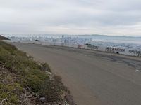 the sky is overcast with a big city in the distance as people walk on the side of a long hill