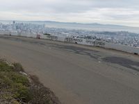 the sky is overcast with a big city in the distance as people walk on the side of a long hill