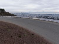 the sky is overcast with a big city in the distance as people walk on the side of a long hill