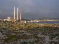 two factory chimneys sit on top of a grassy hill overlooking the ocean and city skyline