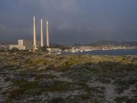 two factory chimneys sit on top of a grassy hill overlooking the ocean and city skyline