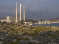 two factory chimneys sit on top of a grassy hill overlooking the ocean and city skyline