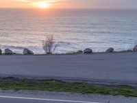 a sunset over the ocean and a long road by the water, with a couple of large rocks on the edge