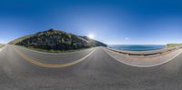 a fish eye view shows the mountains and ocean from a bike path as a mountain sits in the distance