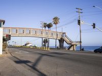 traffic signals hang above an intersection on a beachfront road with palm trees and cars passing underneath it