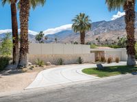 a paved driveway surrounded by palm trees next to a fence and stone wall with mountains