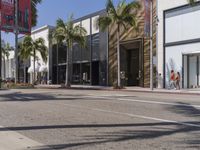 people walking down an empty street by palm trees and an art museum building on a sunny day