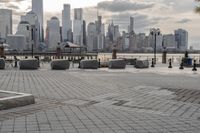 a paved park with benches next to a water body and a skyline of high rises in the background