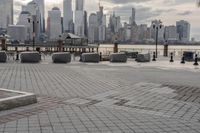a paved park with benches next to a water body and a skyline of high rises in the background
