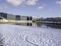 a park bench on a path by the water in snow outside a big city building