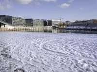 a park bench on a path by the water in snow outside a big city building