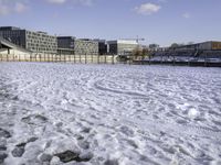 a park bench on a path by the water in snow outside a big city building