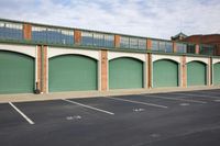 a parking lot has a row of green garages in it and two buildings in the background